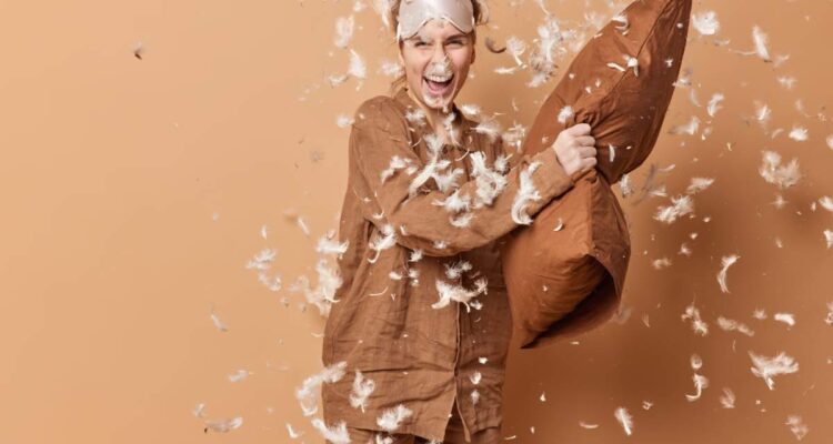 Positive young pretty European woman has pillow fight after awakening wears pajama and sleepmask laughs joyfully poses against brown background with feathers flying around has fun after awakening